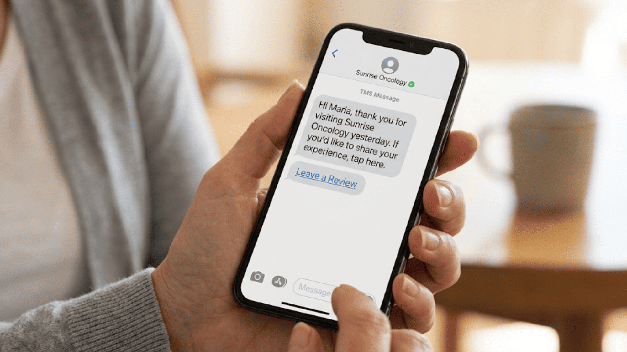 Close-up of a patient's hands holding a smartphone displaying a post-visit review request text from an oncology practice