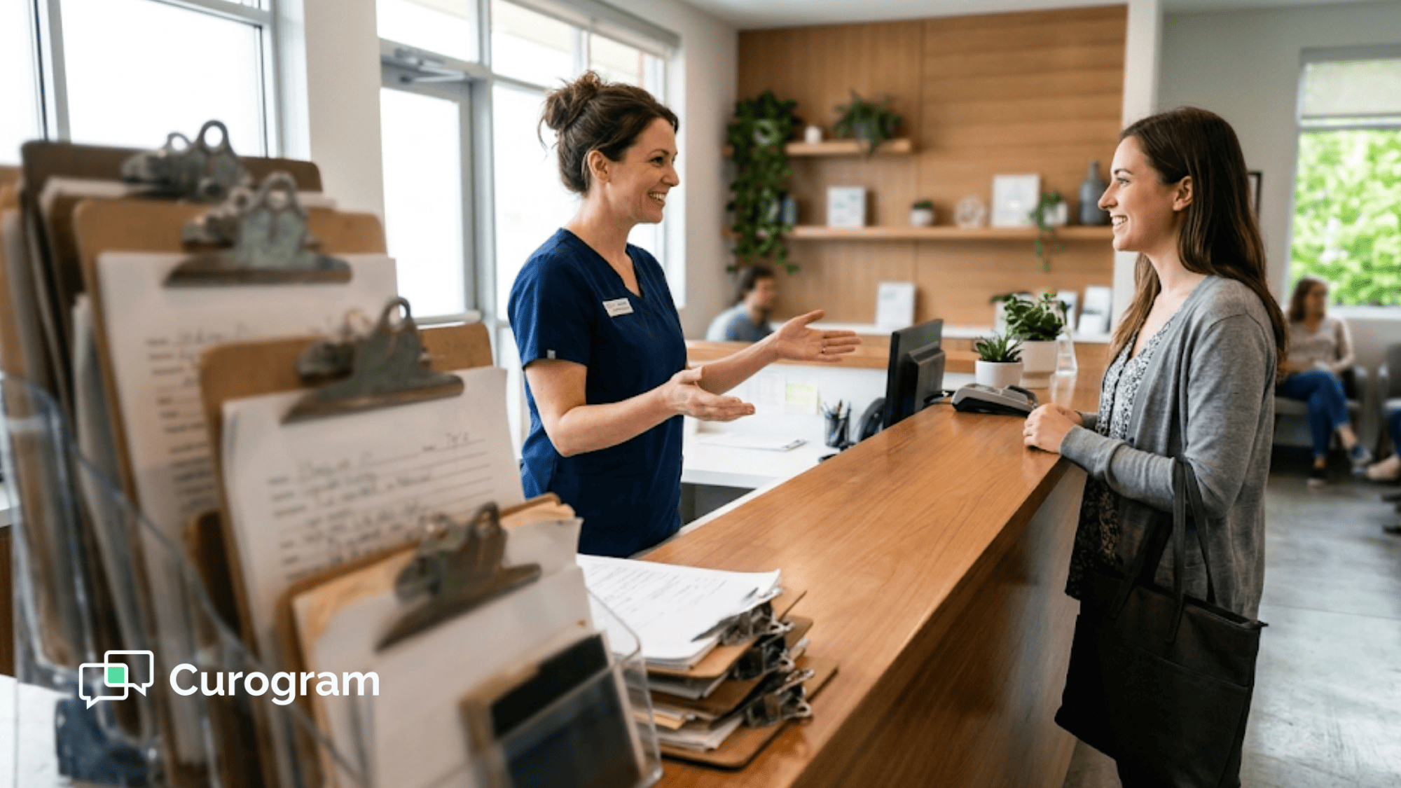 Front-desk staff greeting a patient beside a stack of paper intake clipboards at a clinic reception