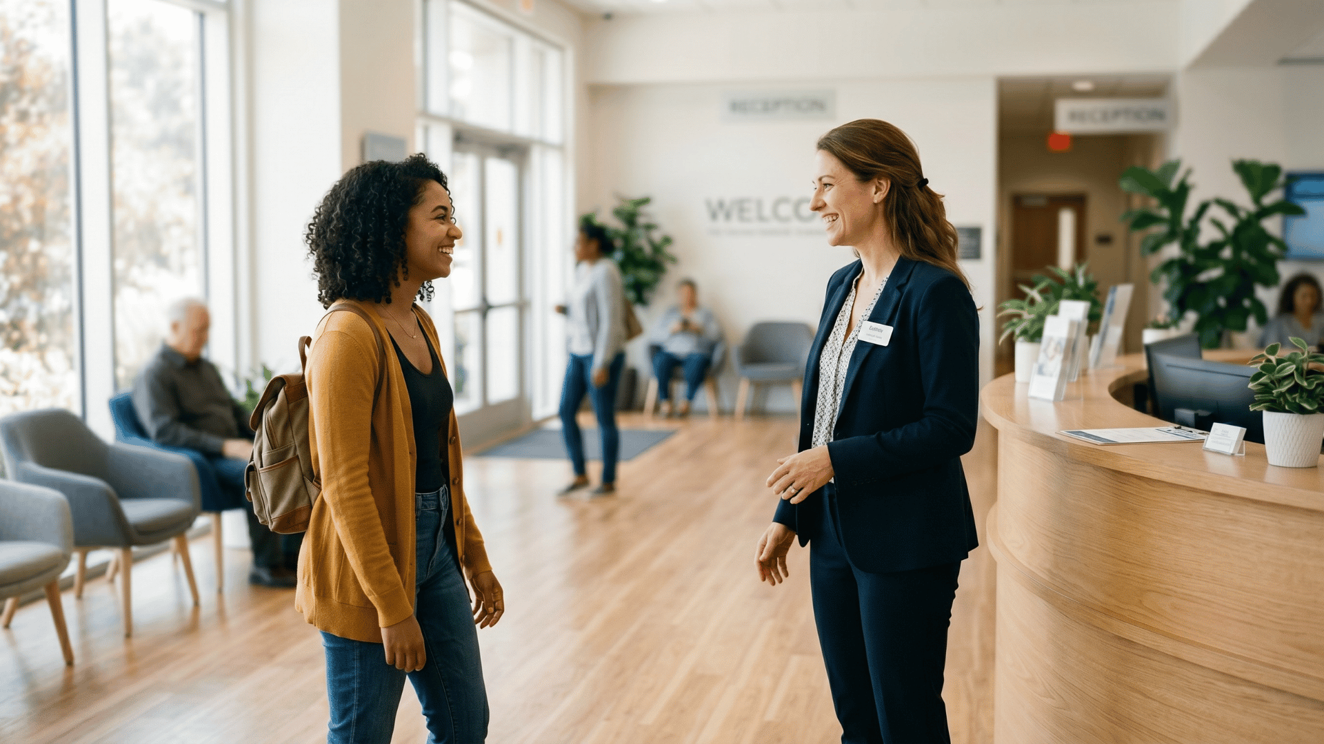 Front desk coordinator standing to welcome an arriving patient in a bright clinic lobby