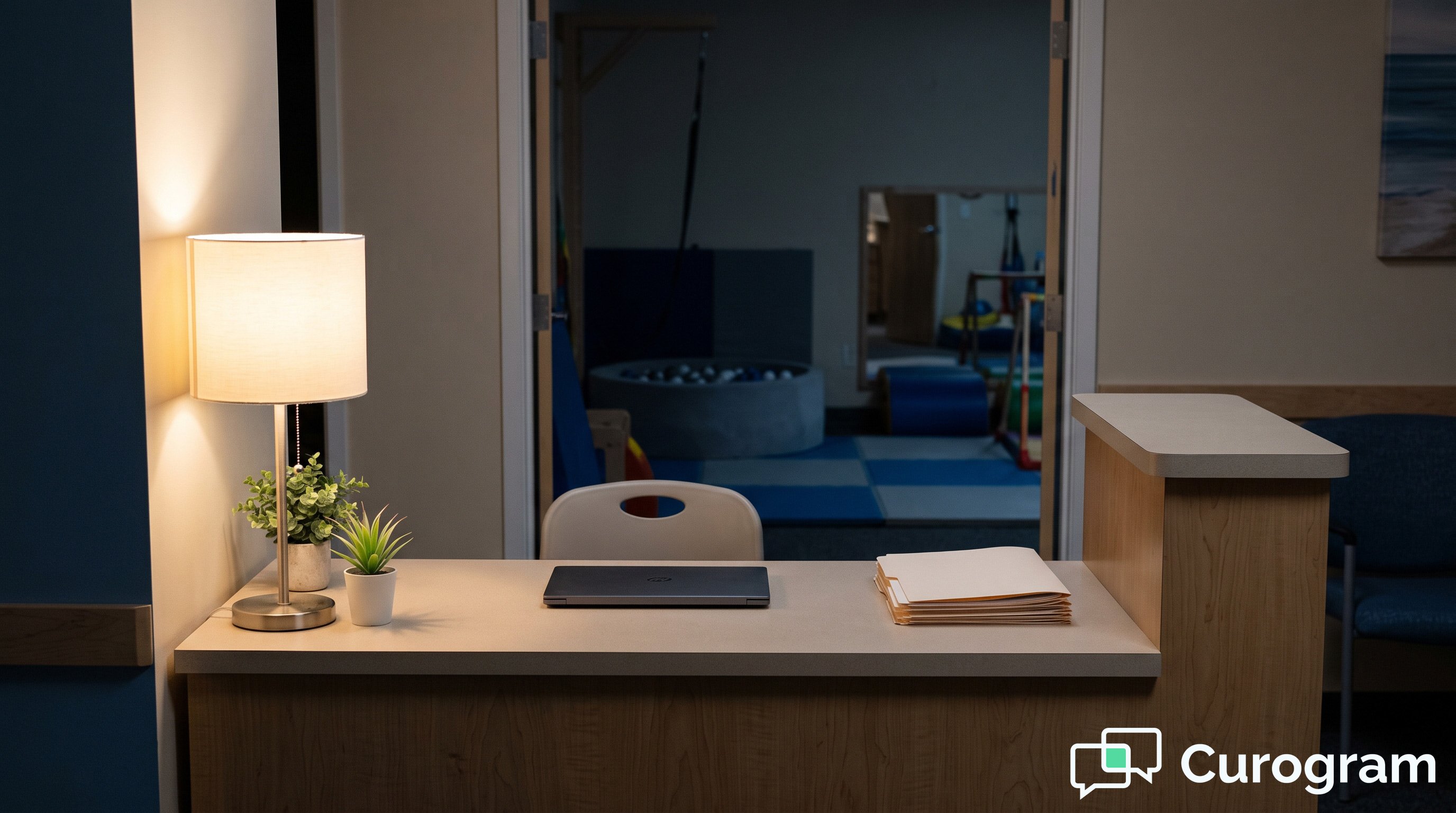 A therapy clinic front desk after hours, with a desk lamp on and an empty pediatric therapy room visible behind it