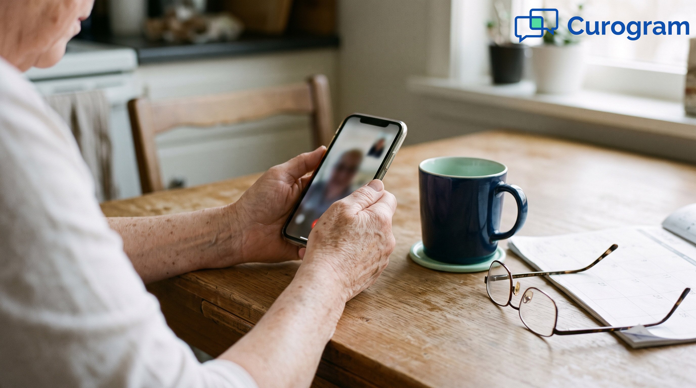 An older person's hands holding a phone showing a video call, beside a mug and a paper calendar